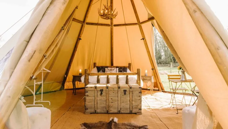 A photograph of the interior of the ultimate tipi, a premium accommodation option at Lost Village. Featuring a double bed, chest and a table and chair set, as well as a clothes rack.